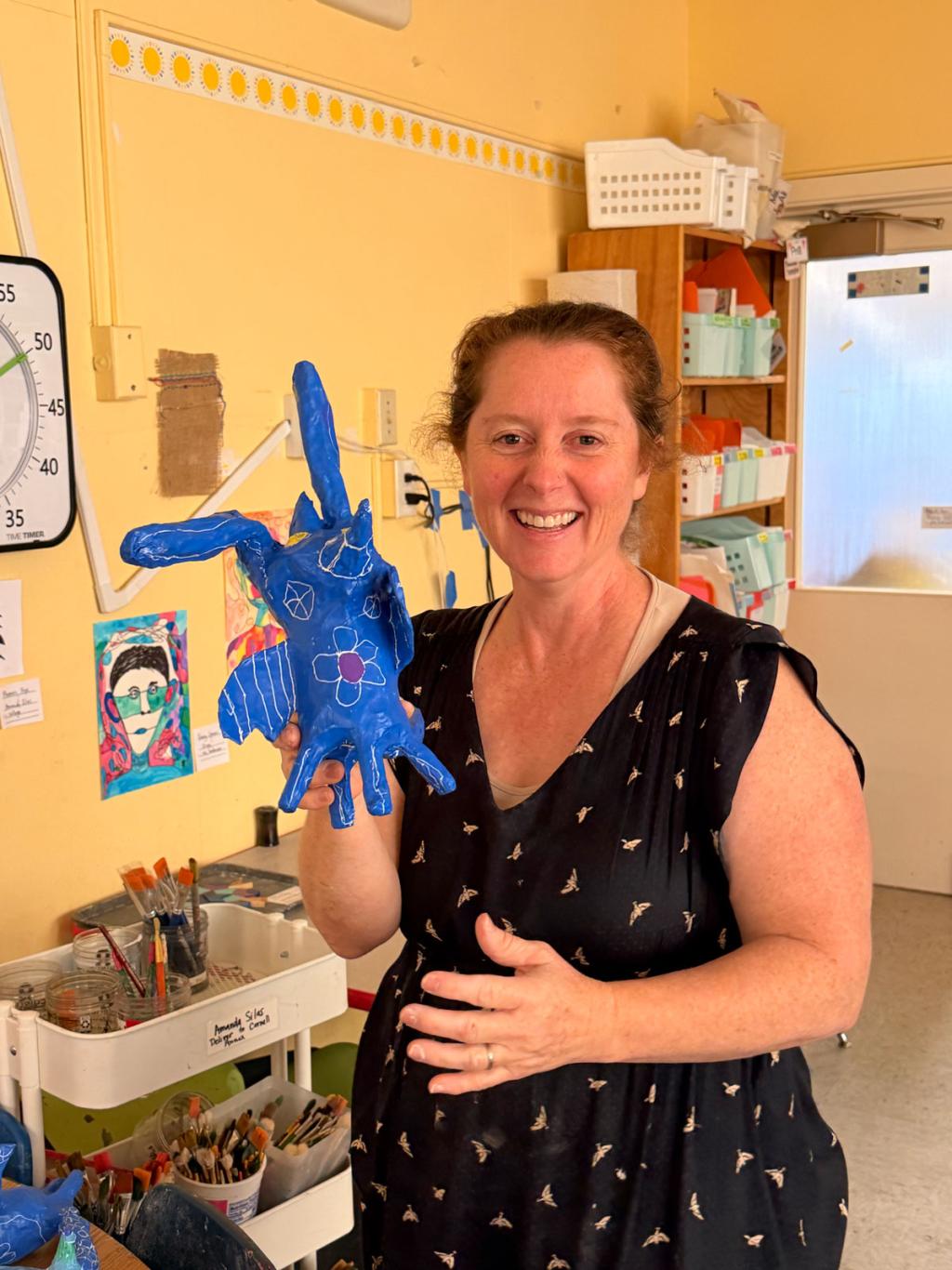 A smiling woman holds up a blue, handmade creature with long ears and wings.