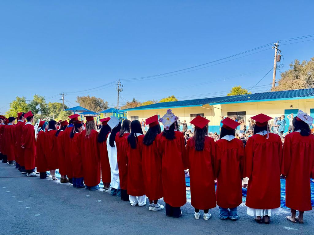 A line of graduates in red gowns and caps stand outdoors, facing away from the camera.