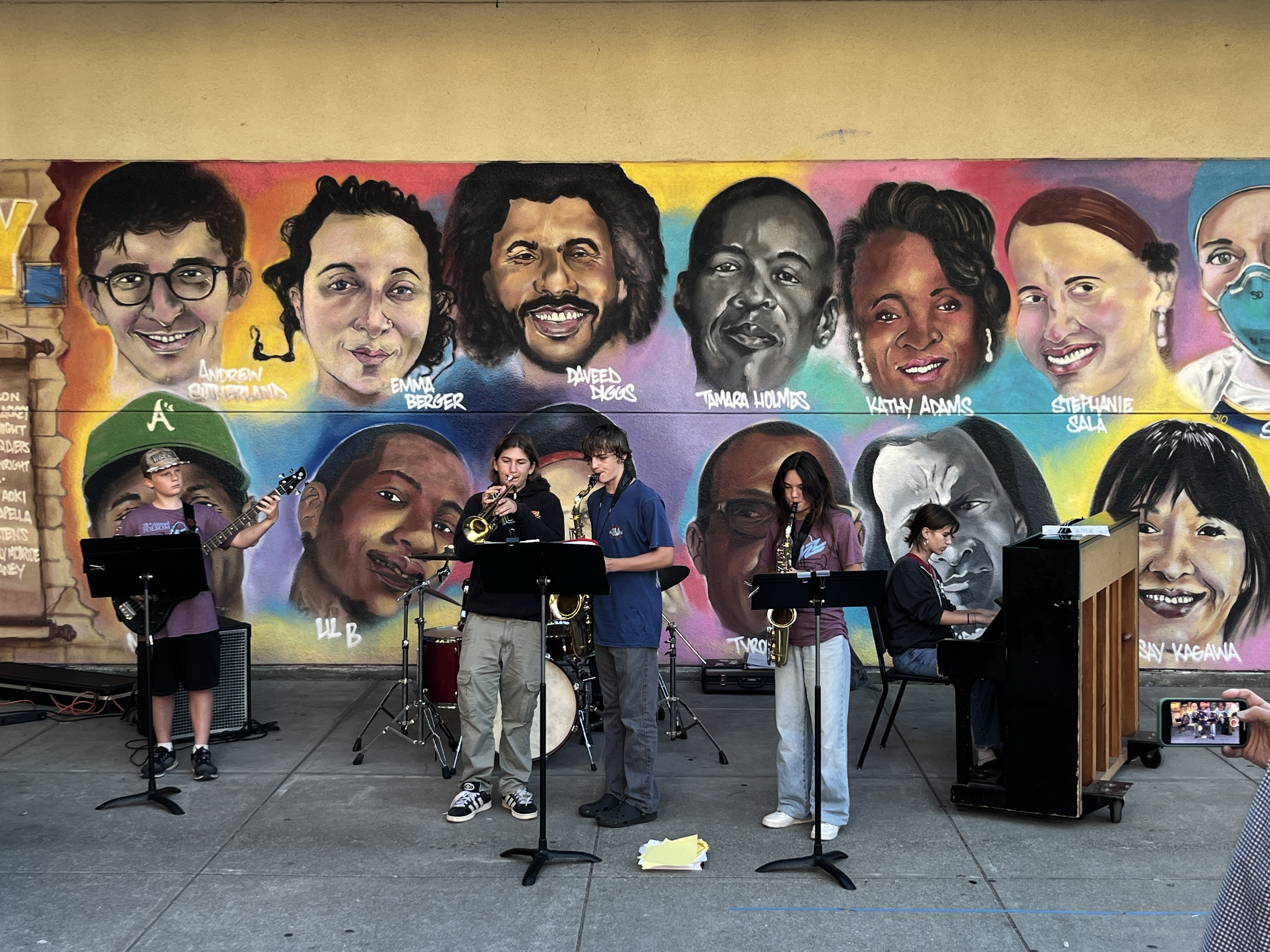 A diverse group of young musicians performs in front of a vibrant mural of portraits.