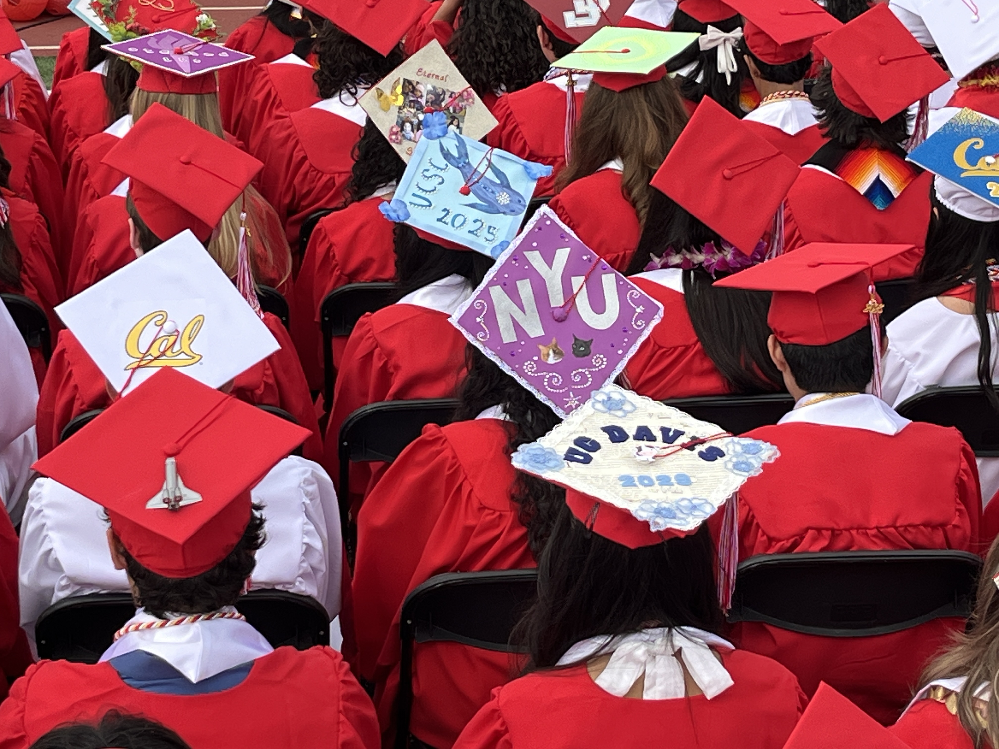 Graduates in red gowns and caps sit in rows, some caps decorated with university logos and years.