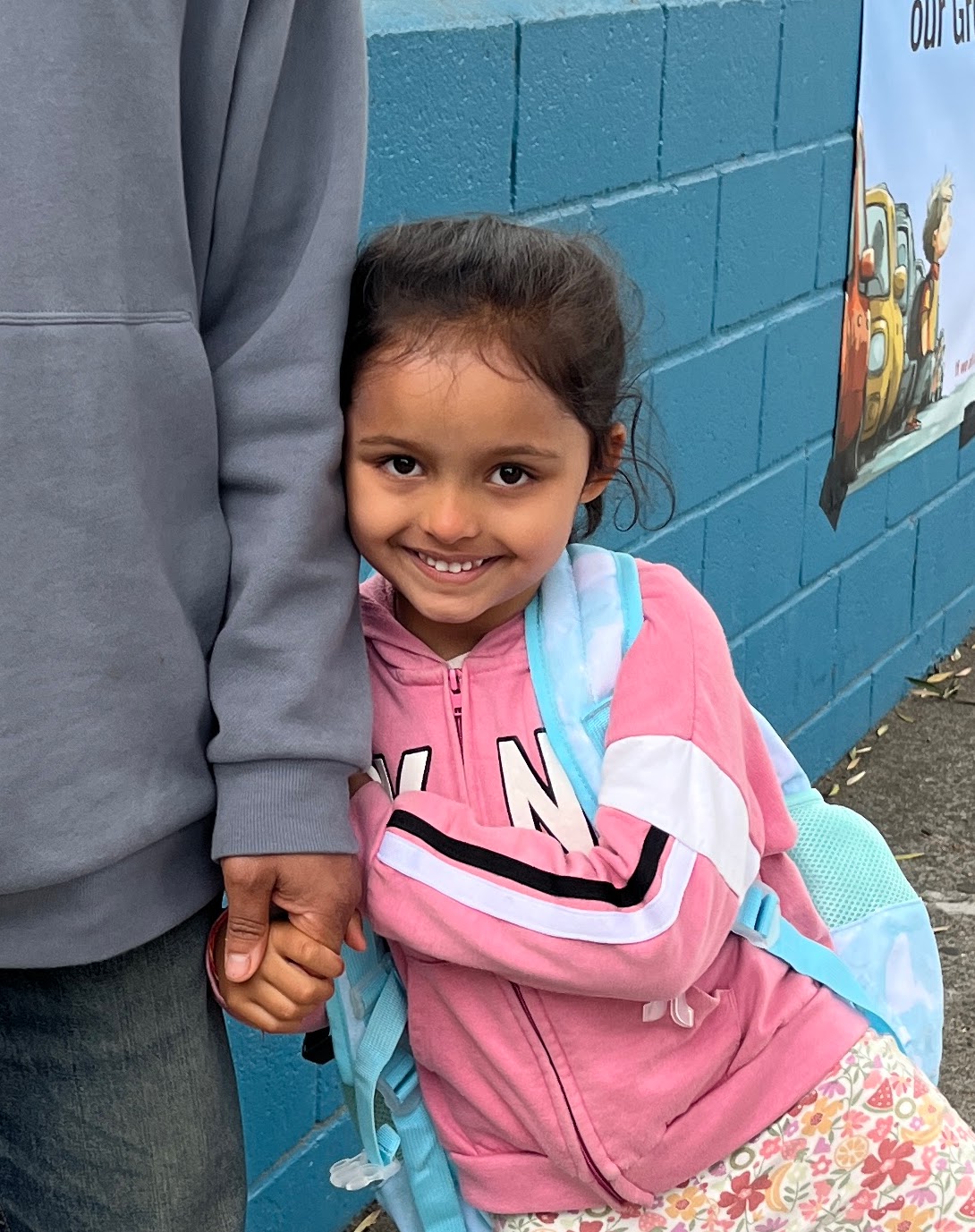 A young girl with a backpack smiles while holding an adult's hand near a blue wall.