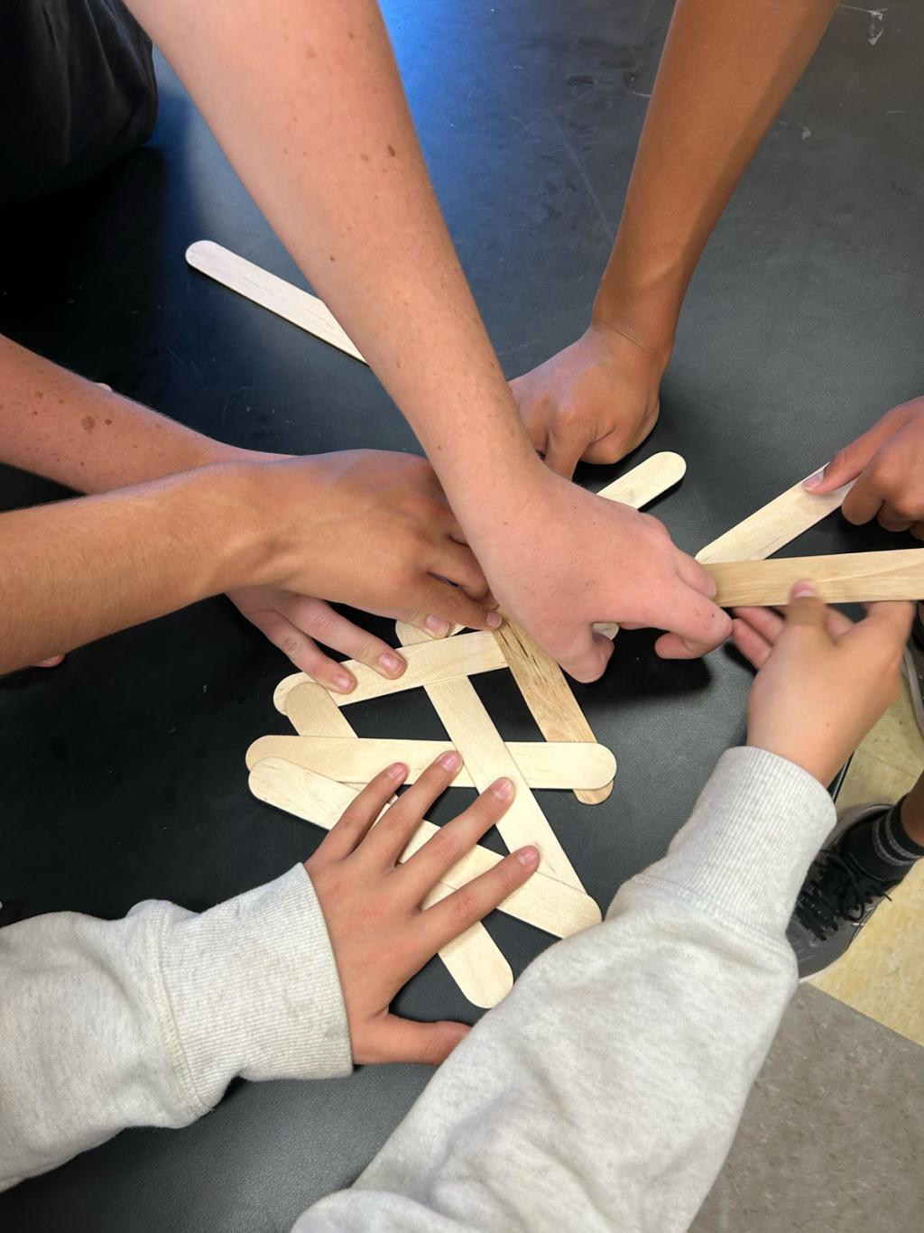 Several hands work together to build a structure with wooden craft sticks.