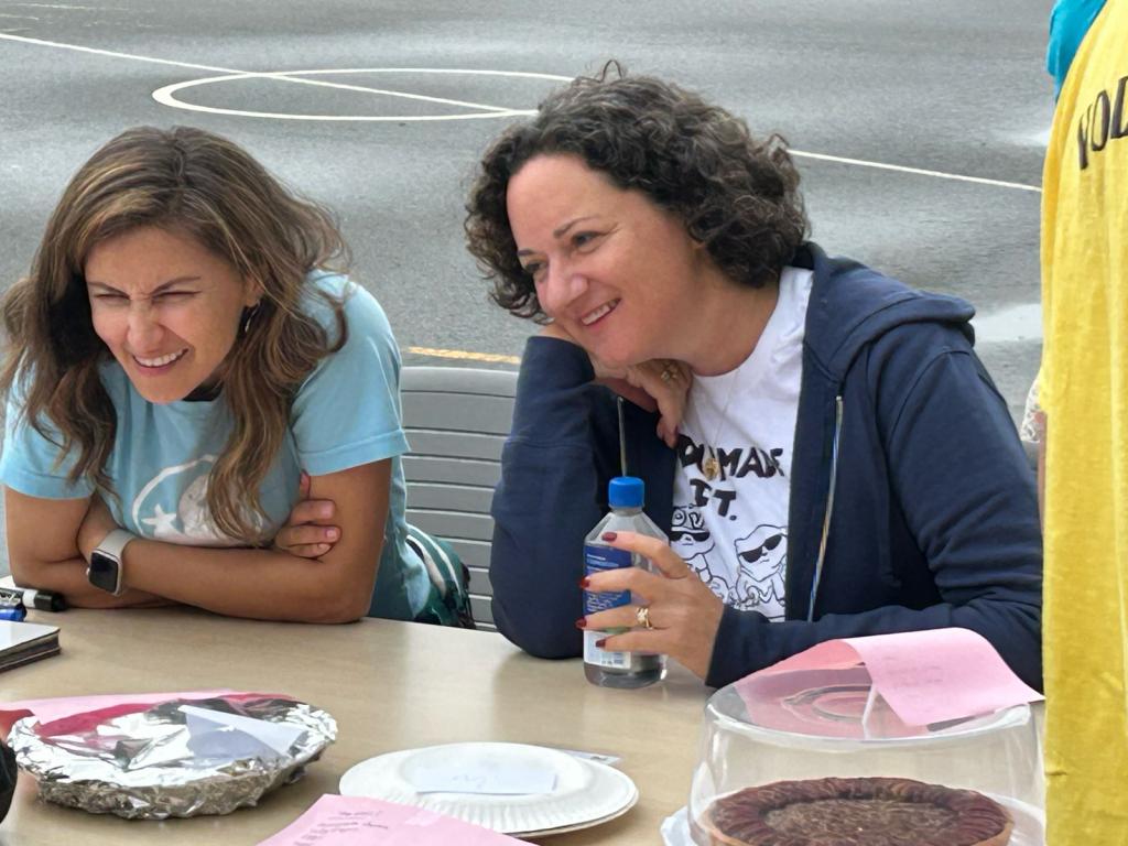 Two women smile and laugh while sitting at a table with food.
