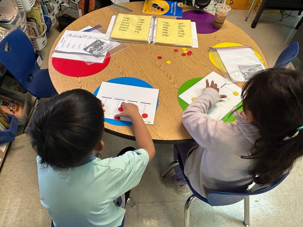 Two children are seated at a round table, engaged in an art or writing activity.