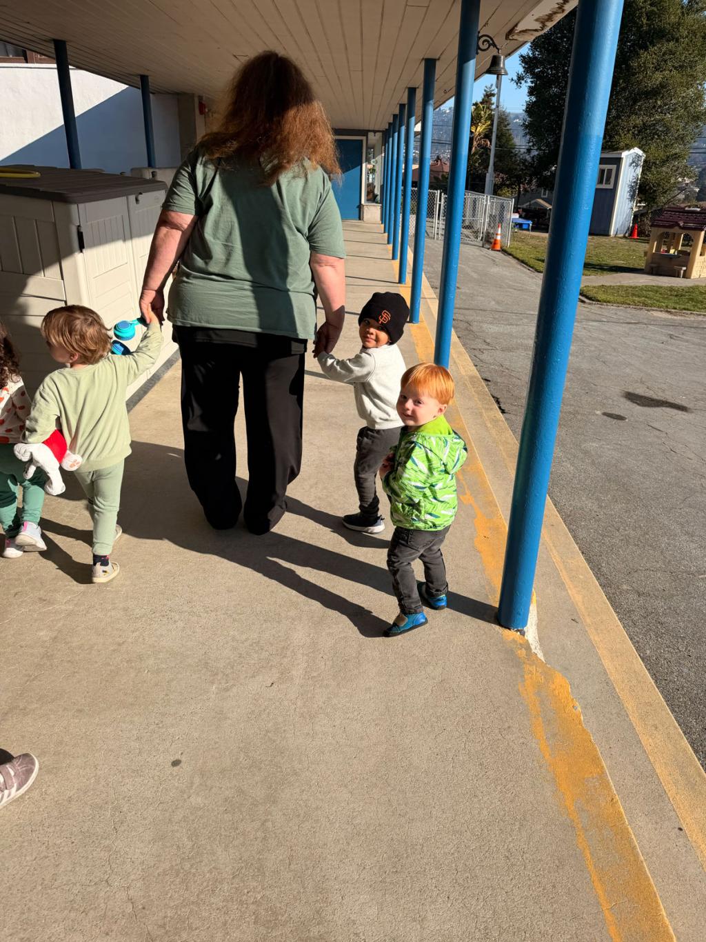 A woman holds hands with two young children walking down a sunny, covered walkway.