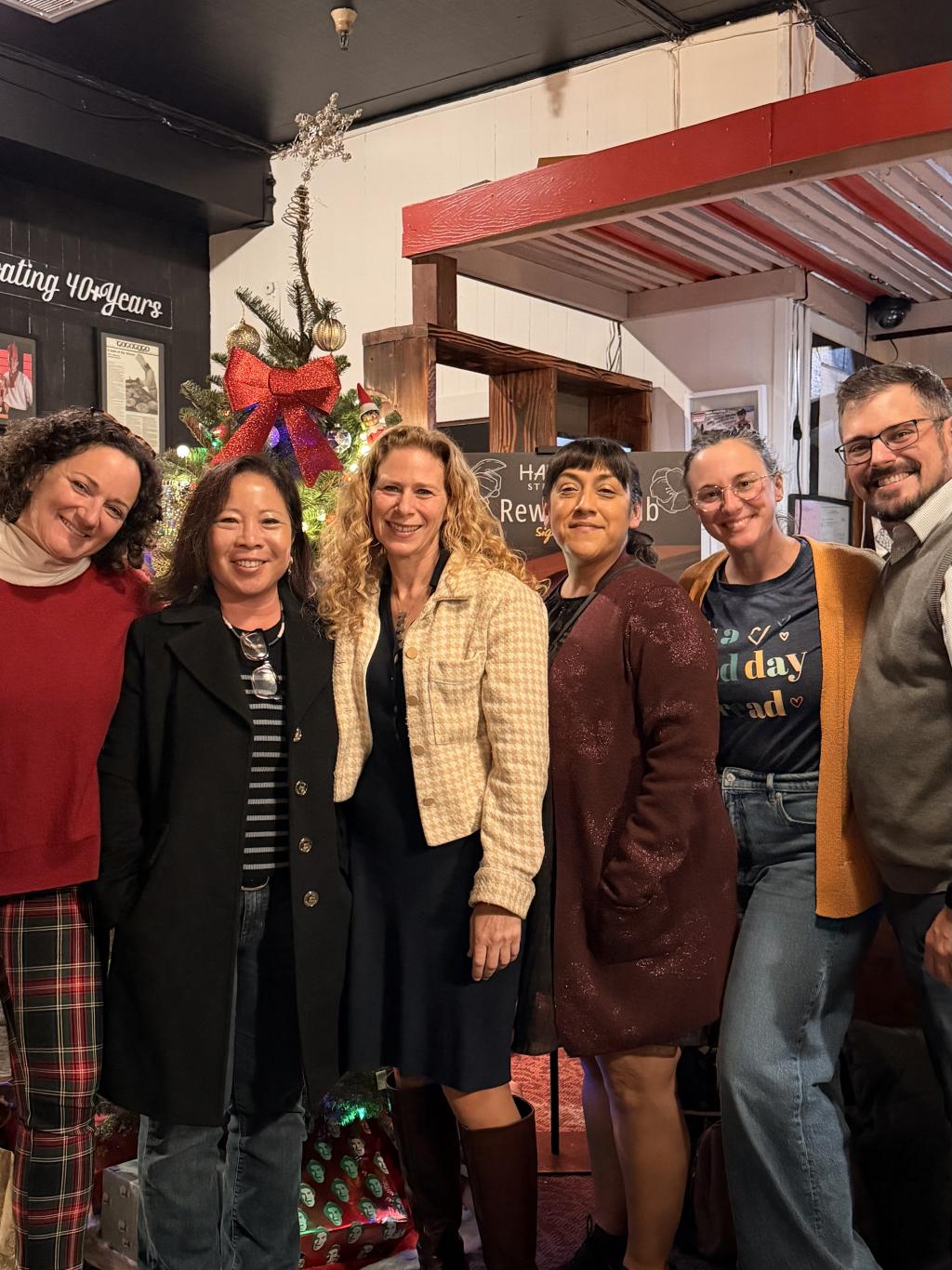 A group of six smiling people stand together in front of a decorated Christmas tree.