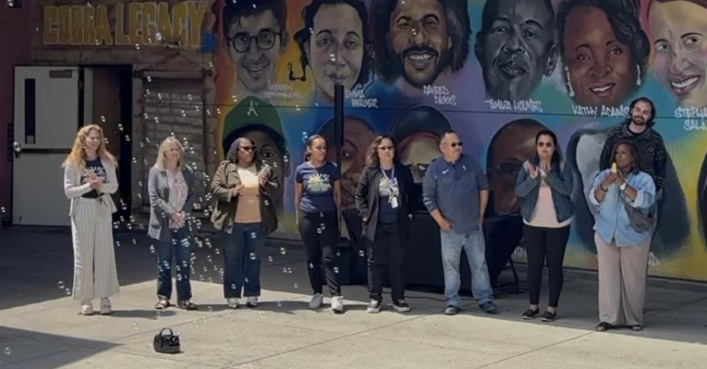 A diverse group of people stand in front of a colorful mural depicting faces.
