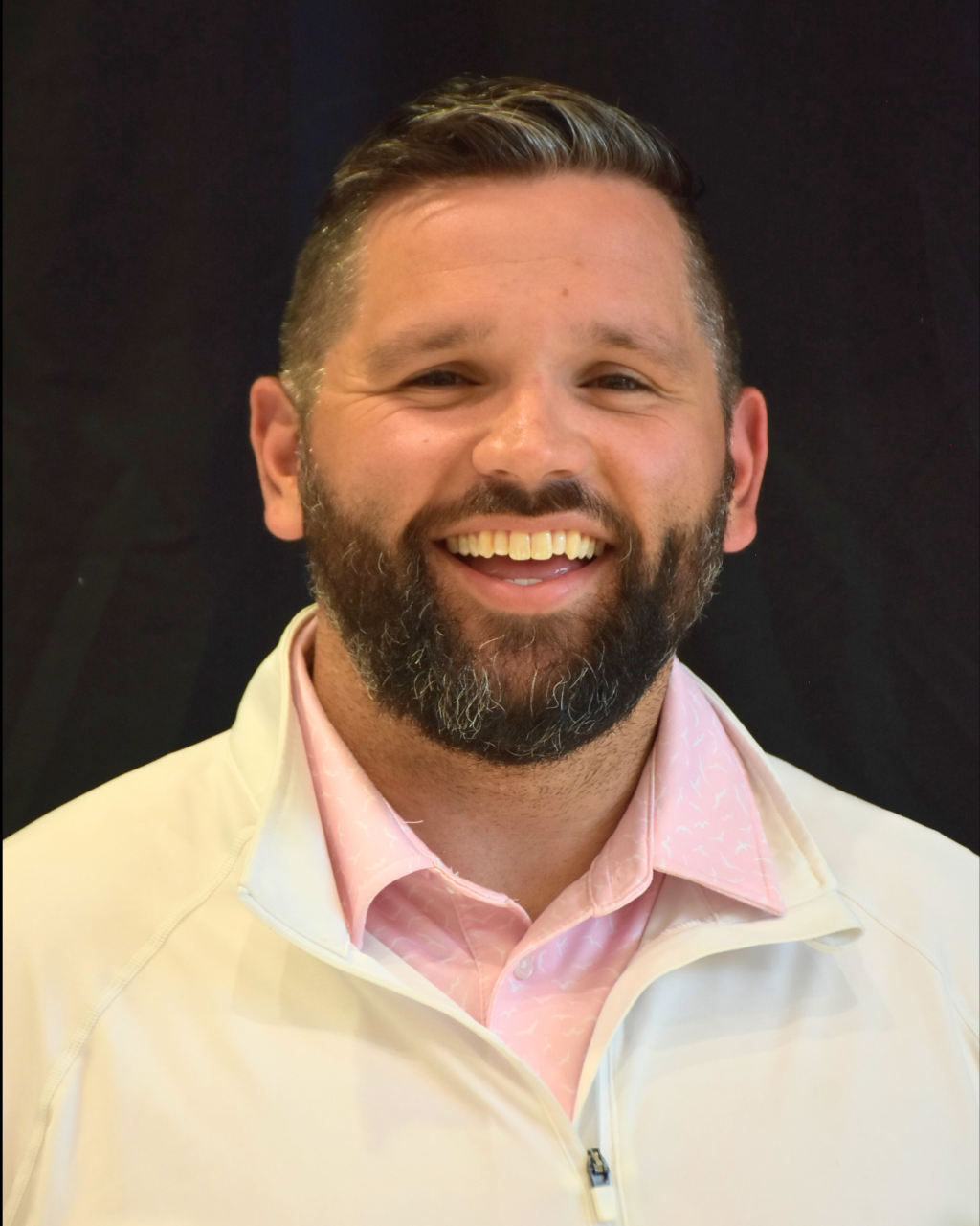 A man with a beard smiles broadly, wearing a pink collared shirt and a white jacket.