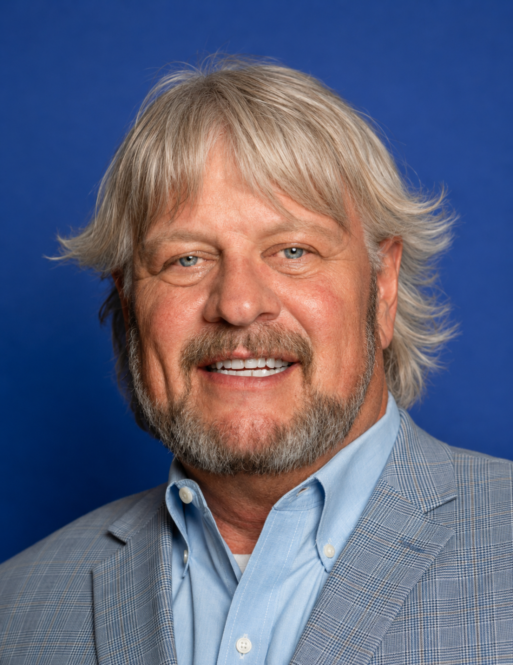 A man with graying hair and a beard smiles at the camera, wearing a light blue shirt and a patterned blazer.