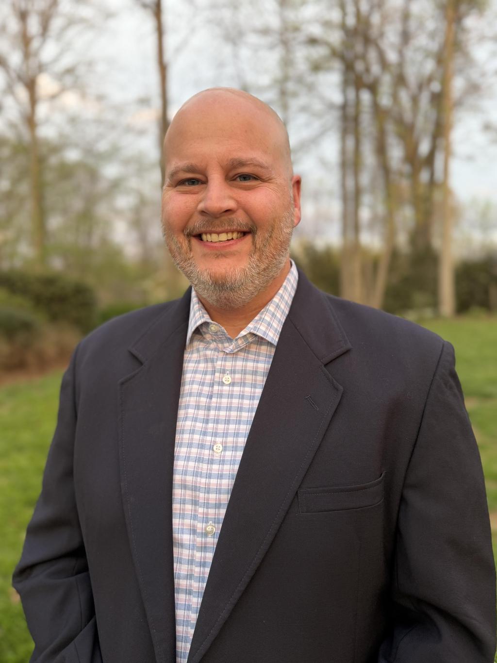 A smiling man in a suit jacket and checkered shirt stands outdoors with trees in the background.