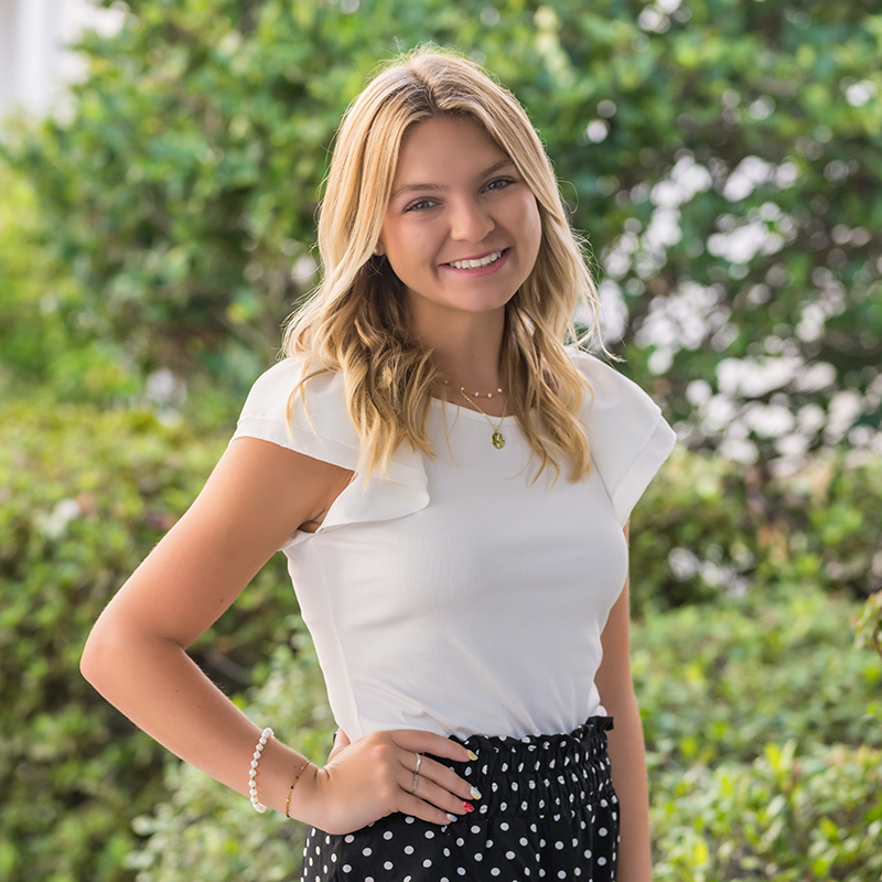 A young woman with blonde hair smiles at the camera, standing outdoors with greenery in the background.