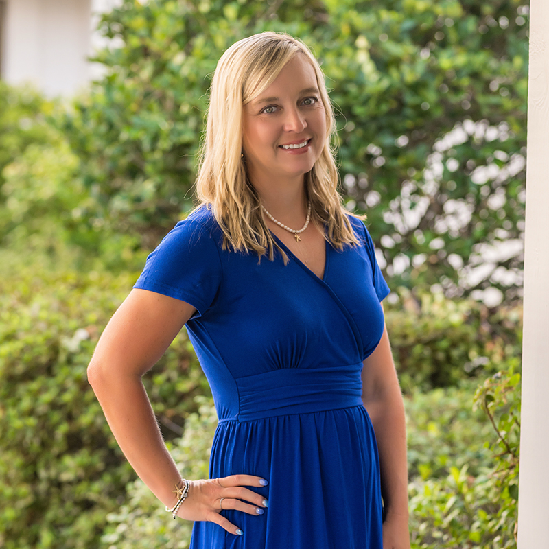 A woman with blonde hair smiles, wearing a royal blue dress and standing outdoors.