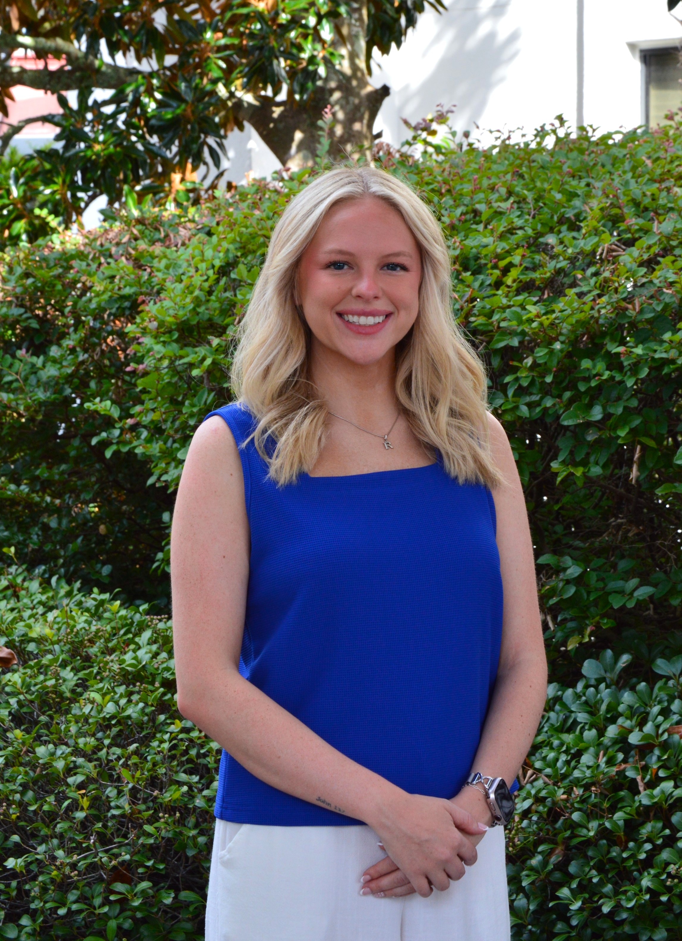 A young woman with blonde hair smiles at the camera, wearing a blue sleeveless top and white pants.