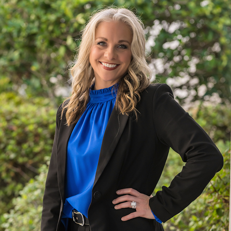 A blonde woman smiles while wearing a bright blue top and a black blazer outdoors.