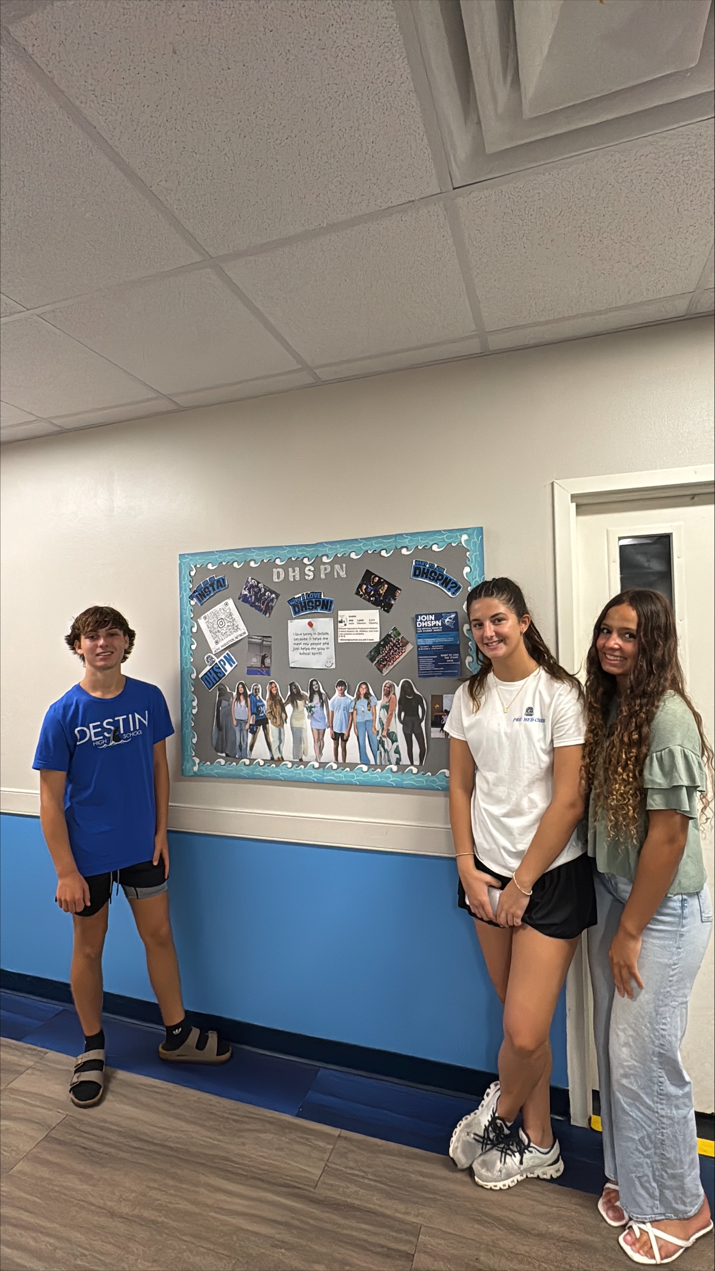 Three young people stand near a bulletin board in a hallway.