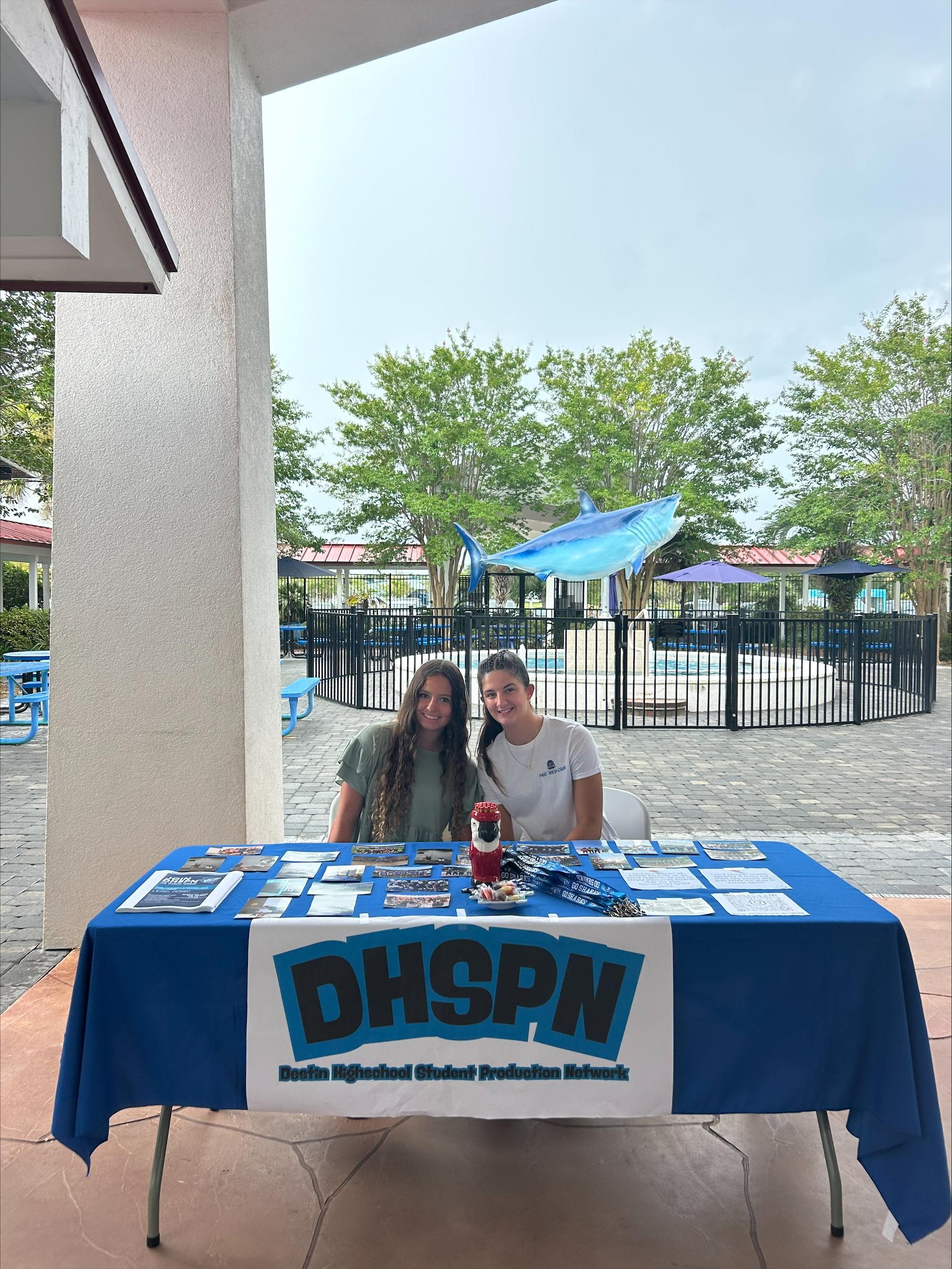 Two young women sit behind a table with informational materials, smiling at the camera.