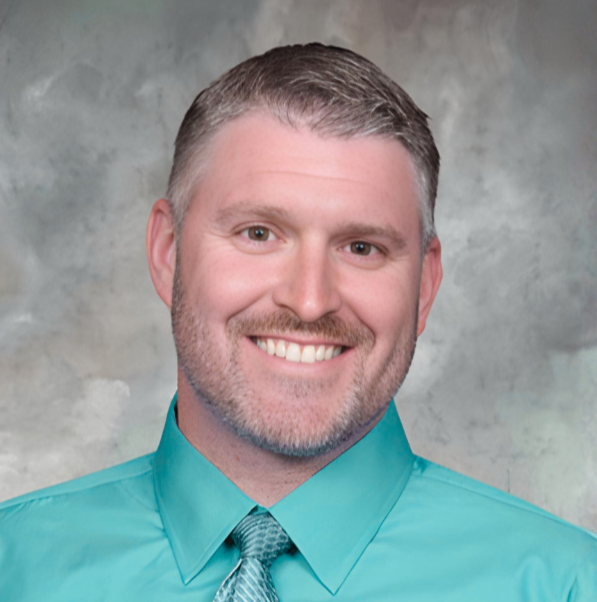 A man with short graying brown hair and a beard smiles warmly, wearing a teal collared shirt and a patterned tie.