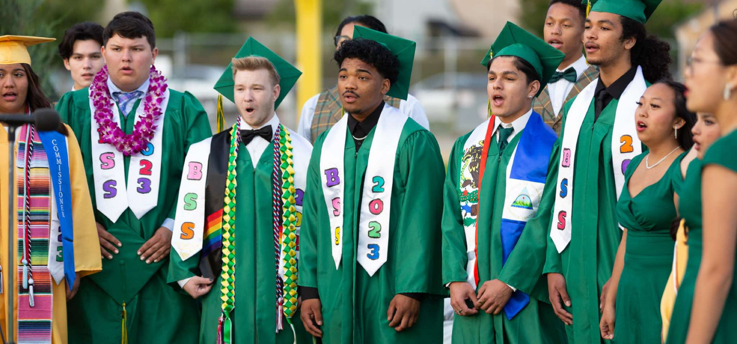 A group of graduates in green gowns and caps stand together, some wearing leis and sashes.