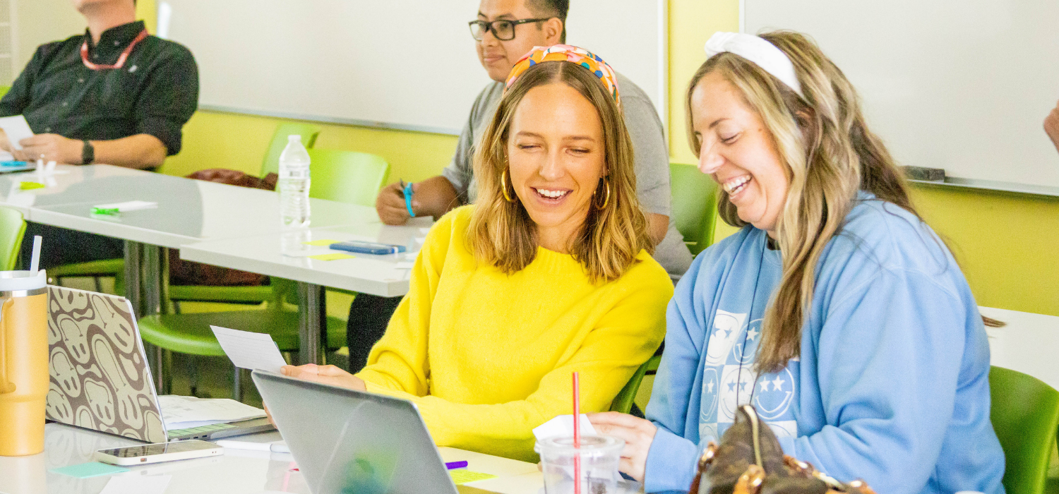 Two women smile while looking at a laptop in a bright, modern classroom.