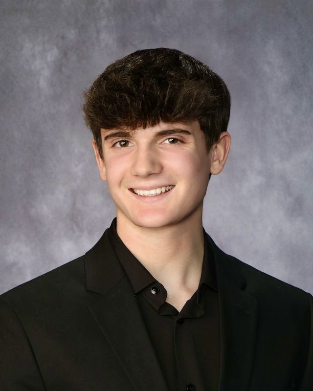 A young man with dark, curly hair smiles confidently, wearing a black suit jacket over a black collared shirt.