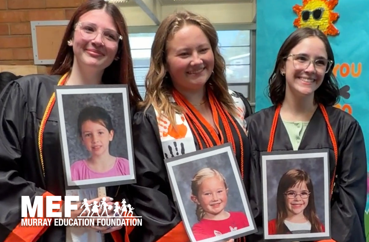 Three smiling young women in graduation gowns hold up framed photos of themselves as children.
