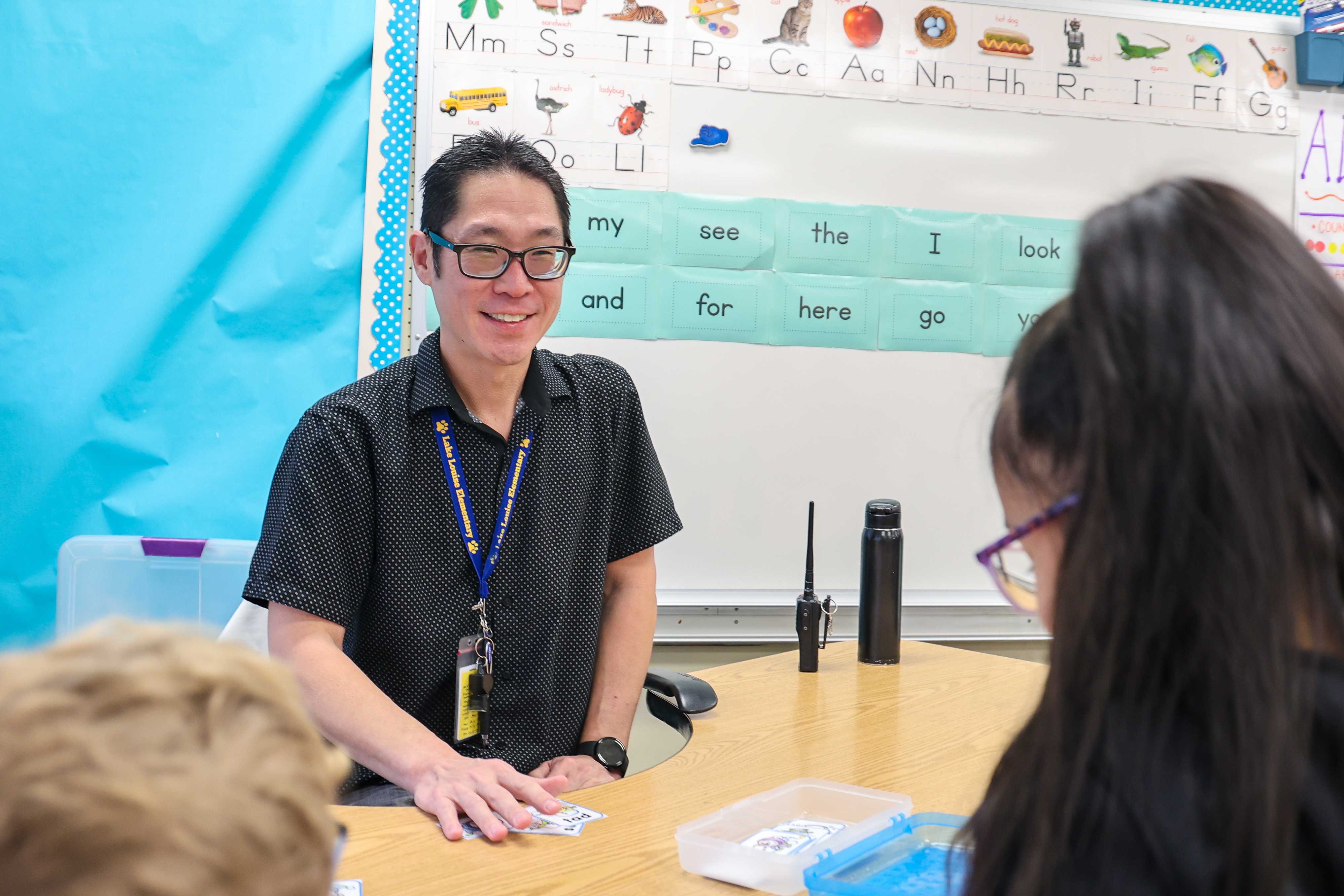 A teacher smiles while speaking to students in a classroom setting.