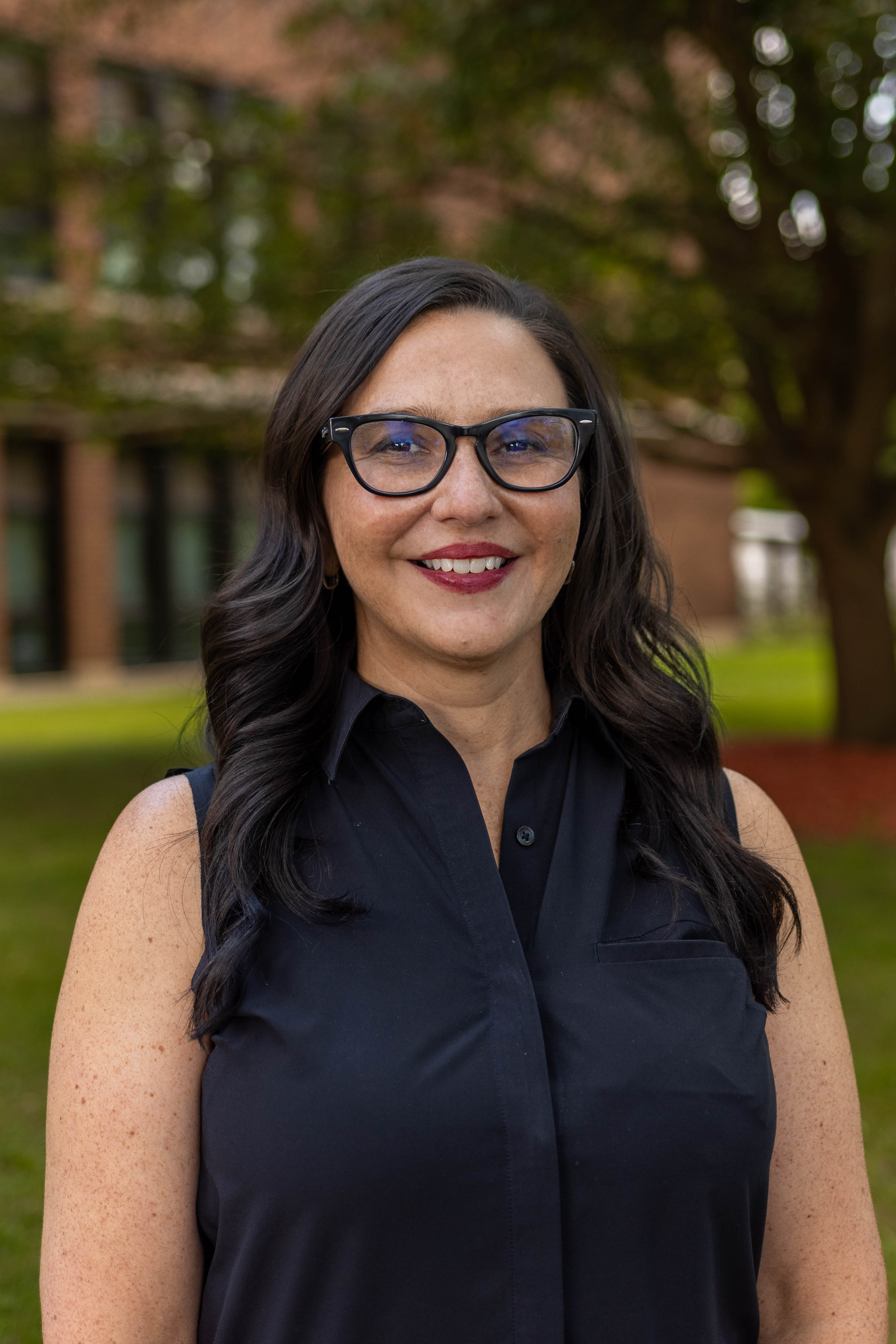 A woman with dark hair and glasses smiles at the camera outdoors.