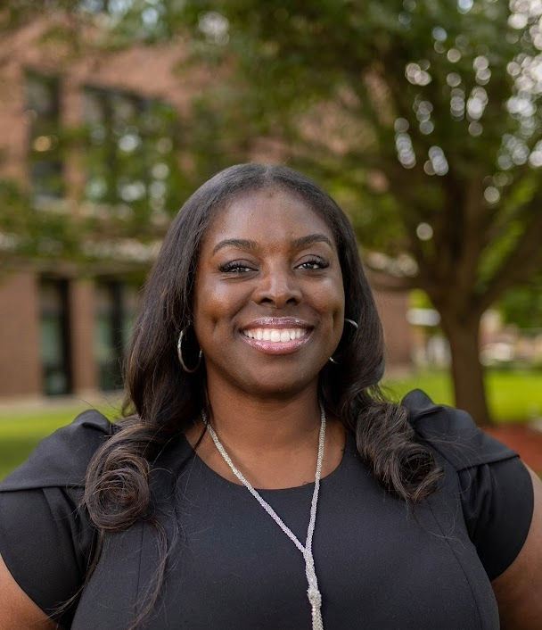 A woman with a warm smile poses outdoors, wearing a black dress and necklace.