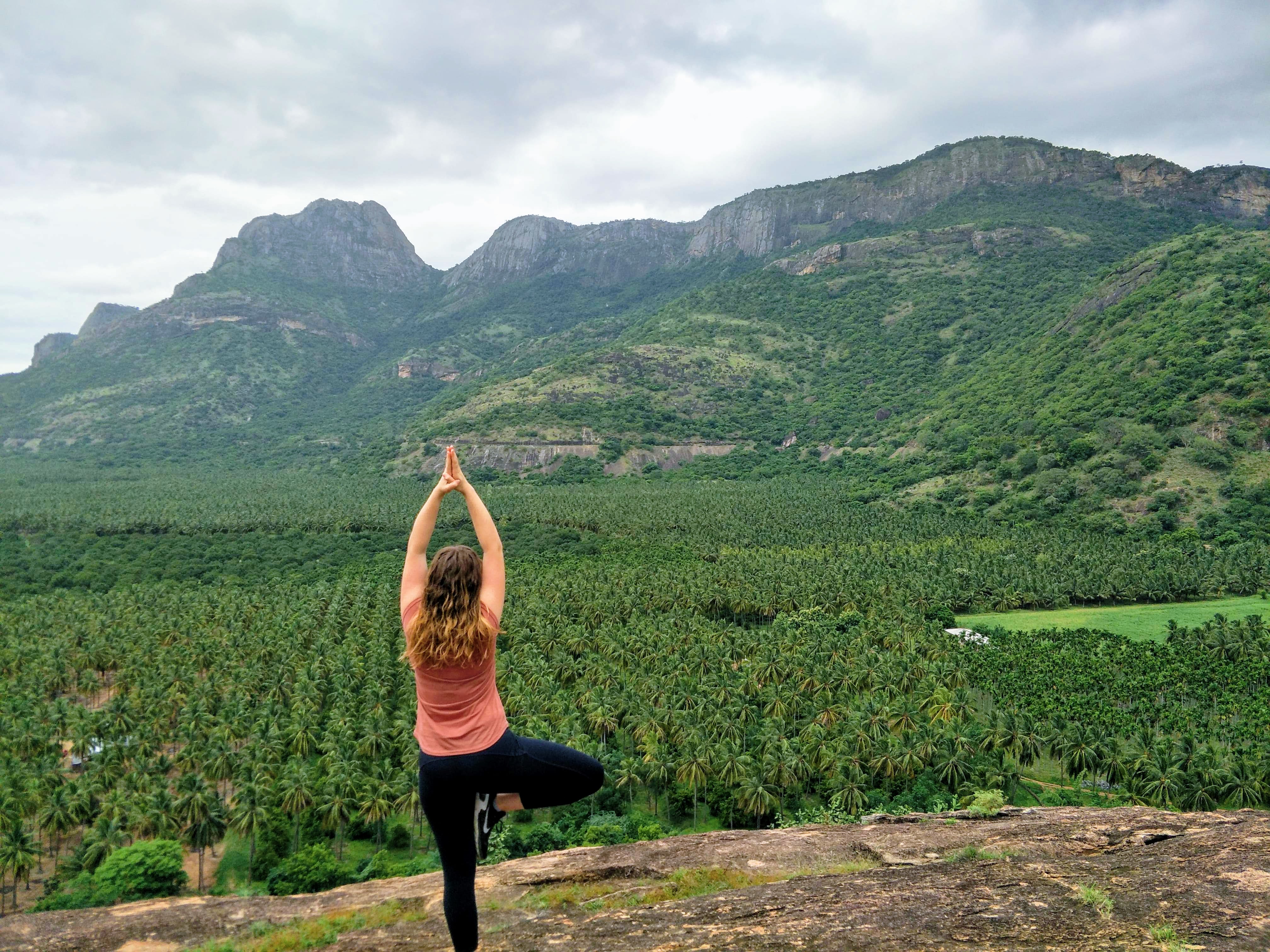 Woman on a rock with a view of many banana trees in trees below and another rock that appears like a woman sleeping in the distance. Woman is standing in tree pose, a yoga position. 