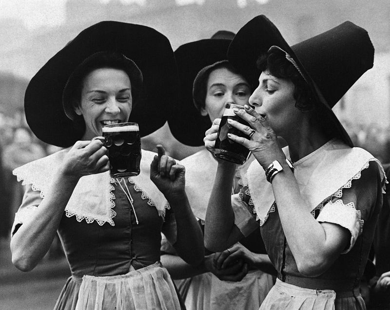 a black and whit picture of a group of womans drinking dark beer in witch like clothes