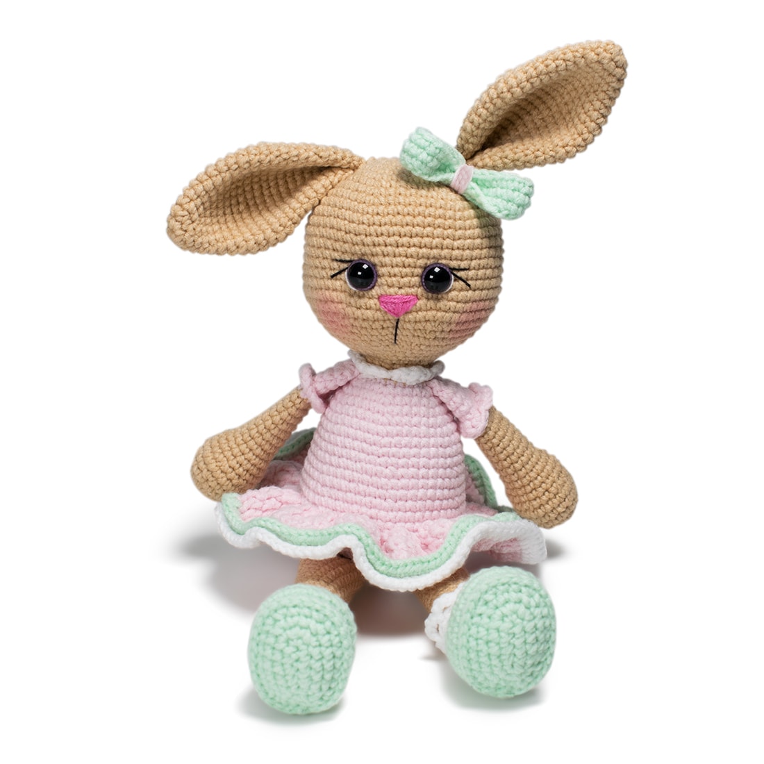 A crochet bunny wears a pink dress, a green bow on one ear and green shoes while sitting.