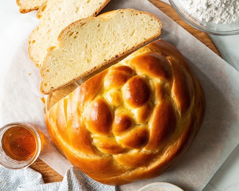 This is an image of Challah, seen from above. It is yellow and golden-brown, with an almost circular shape, and bumps and lumps on top from where the dough was braided before baking. Three slices have been cut off, and a small ramekin of honey sits on a white tablecloth to the left.