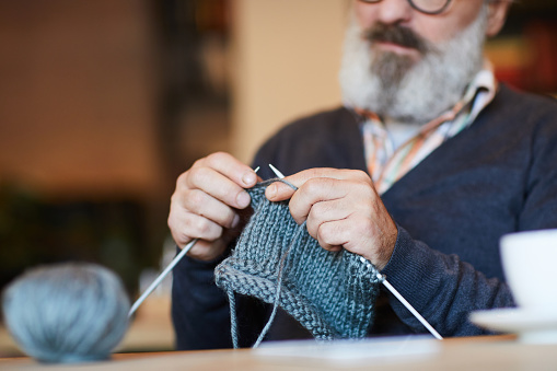 View from the front of a man with a medium white and grey beard knitting. His face is half out of frame and out of focus, 
  bringing his hands with his ice blue knitting project into focus.