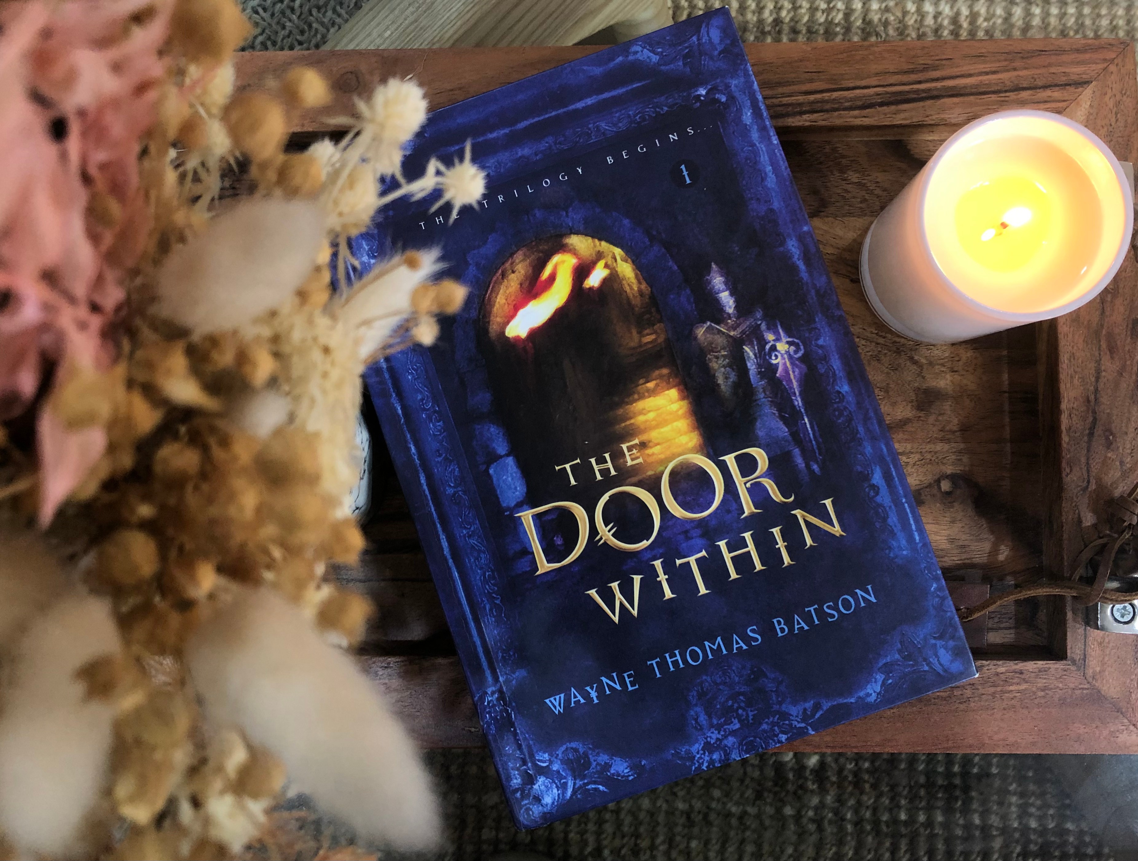 A hardcover copy of the Door Within next to a lit candle and a vase of dried flowers