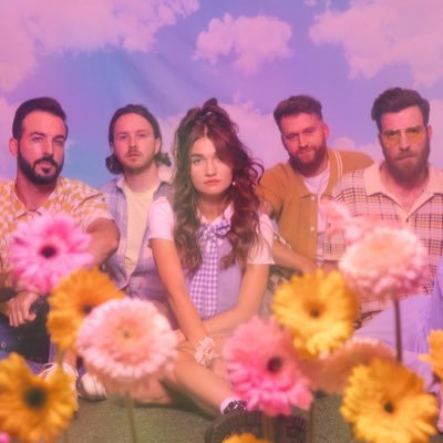 A group of five people sitting. A woman is in the middle with two men on each side. The background is blue with clouds.