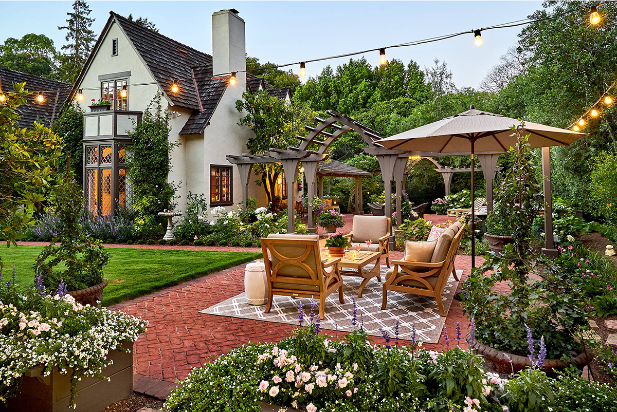 House with garden that has a grass field, paved sitting area with string lights above and flower beds surrounding it.