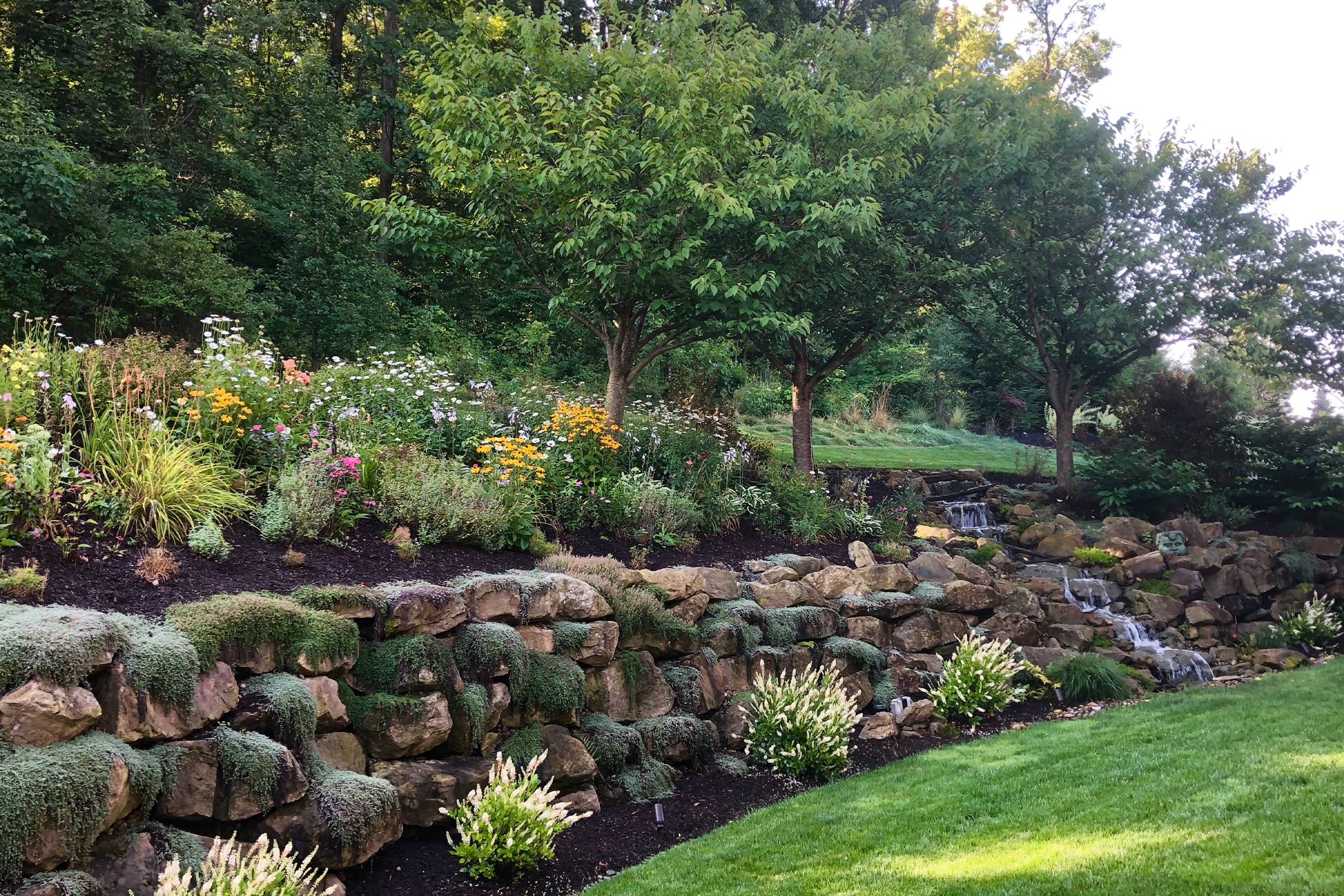 Garden with rock wall with flower bed on top, water cascading down the rocks and a lawn below.