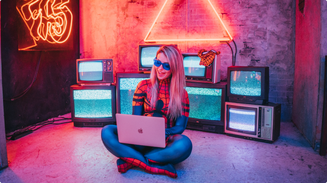 An image of Kelly dressed like spider-man. She is wearing a May Parker Spider-girl suit as she sits on the floor on her pink laptop. She is in a neon light lit room, wearing sunglasses and her blonde hair is straight.