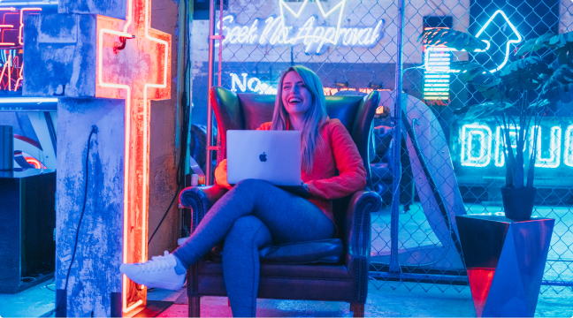 An image of Kelly sitting on a leather chair with her pink laptop. She is laughing. She is sitting next to a neon light cross, in a room full of neon lights.