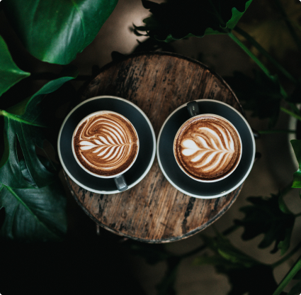 This an image of two artisial lattes and coffees on a wooden table surronded by palm trees.