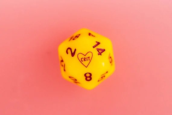 yellow d20 die on a pink background displaying a heart with 'CRIT' written on the 20 side