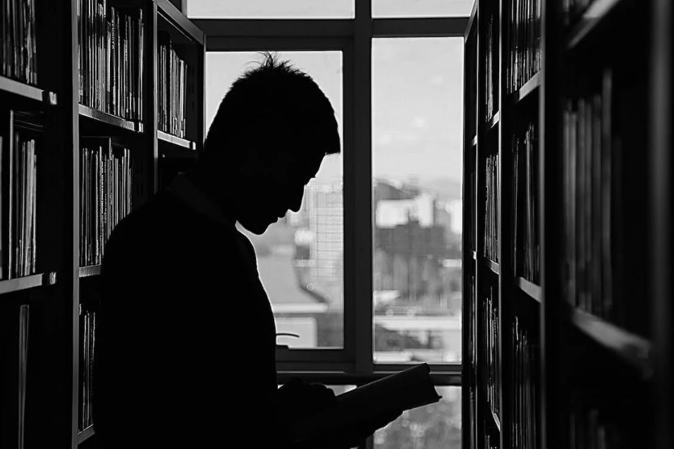 Black and white photo of man reading intently in-between library shelves.