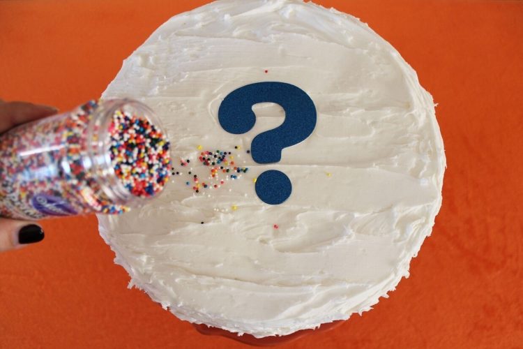 Picture of a white cake with a blue question mark on it and someone shaking sprinkles on top