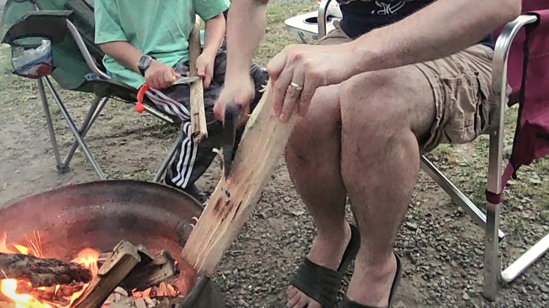 Dad shaving wood with an axe for the campfire and child widdling with a knife