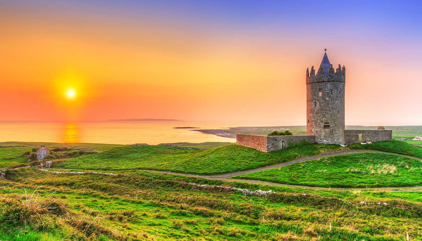 Image of a medieval tower on top of green fields, with a sunset over the sea as background