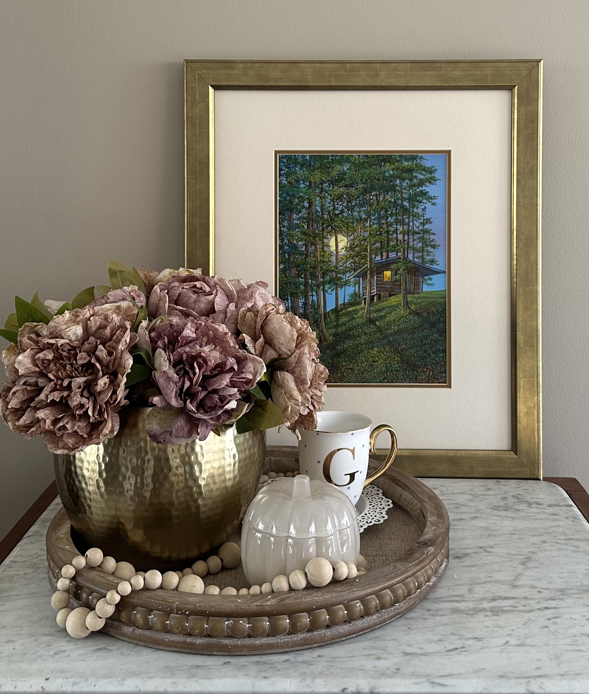 Picture of a marble top table with a tray that has a gold vase filled with light purple flowers, a pumpkin shaped candle and a tea cup with a gold G on it. In the background there is a painting in a gold fram of a hut in the woods.