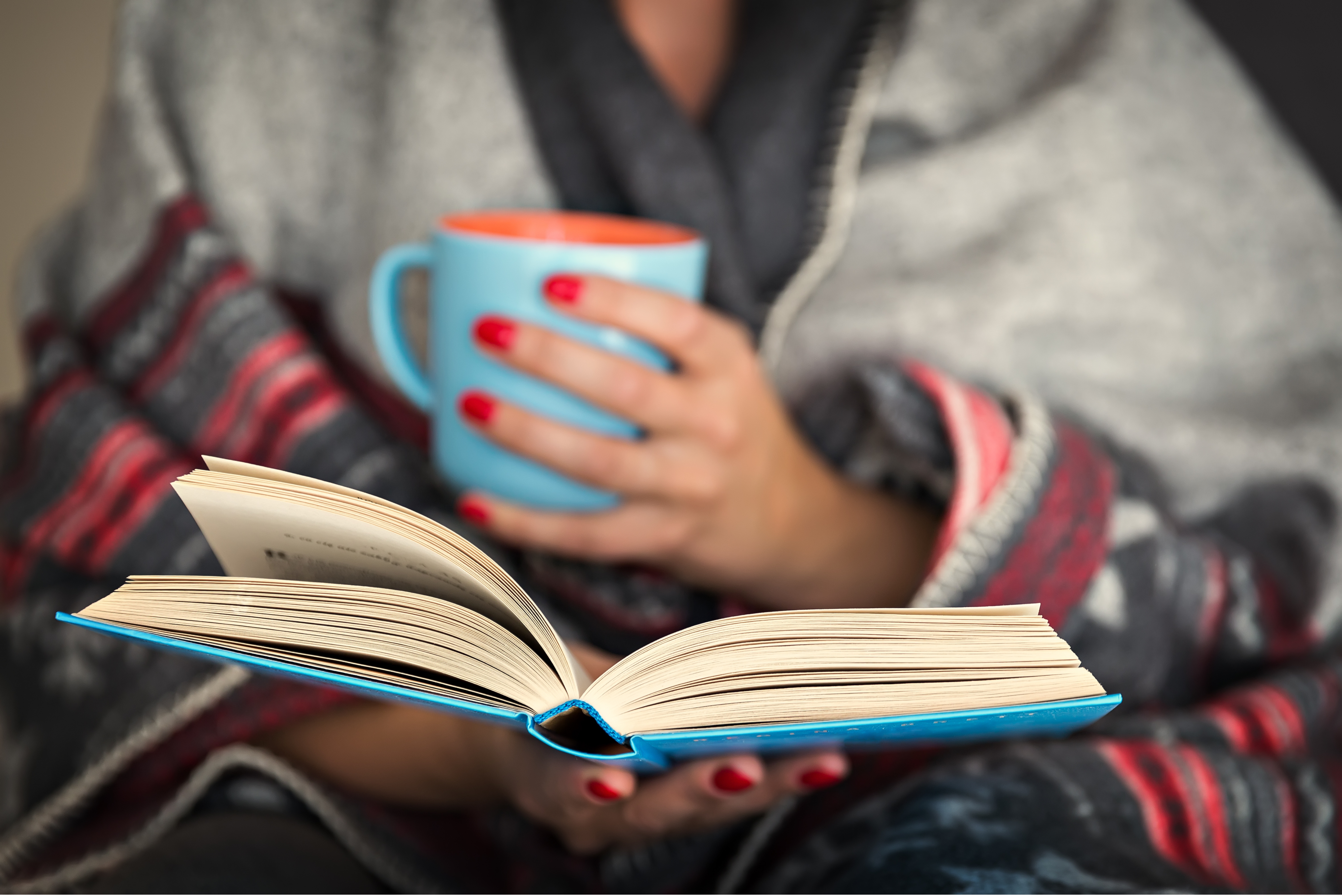 person wrapped in a blanket reading a book and holding a cup of coffee