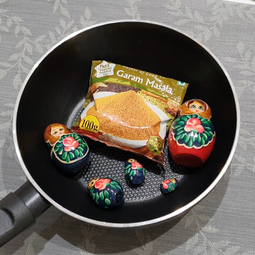 A frying pan containing a bag of garam masala and five dolls