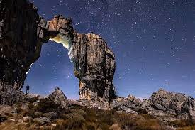 Two hikers walking on a sunlit rocky mountain ridge, one leading and one following, both wearing backpacks; 
         they traverse a narrow trail overlooking a broad green valley under a blue sky with scattered clouds. No visible text in the image.
          Tone calm and adventurous.