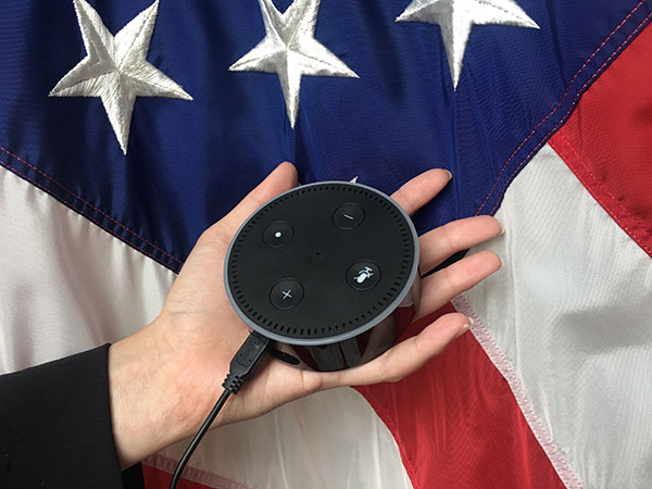 A hand holds an Echo Dot from Amazon in front of an American flag.